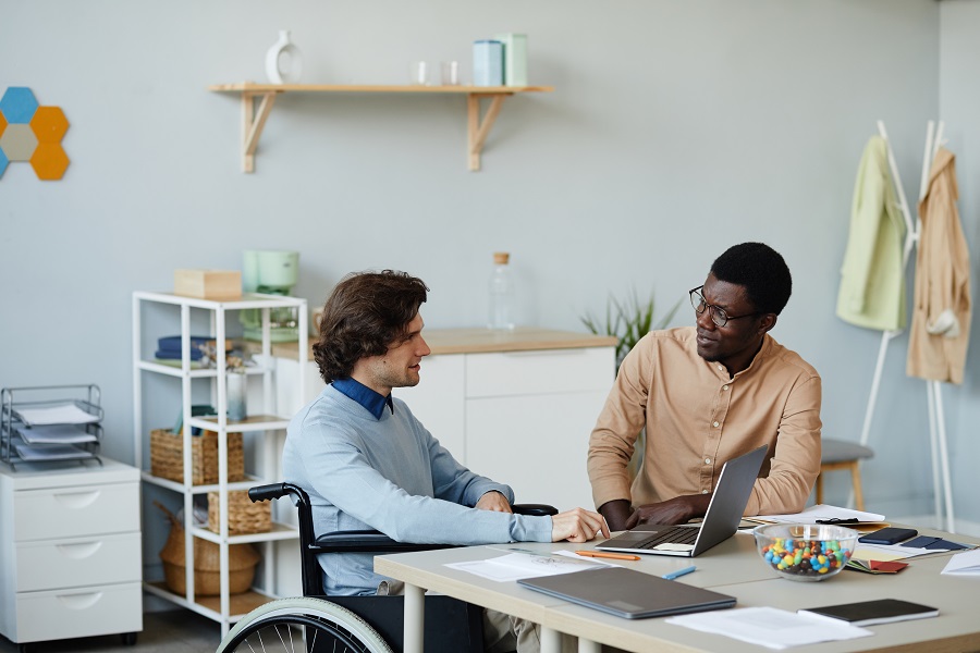 Ritratto di un giovane con disabilità che parla con un collega sul posto di lavoro in un ambiente di ufficio minimo, concetto di opportunità di lavoro accessibile